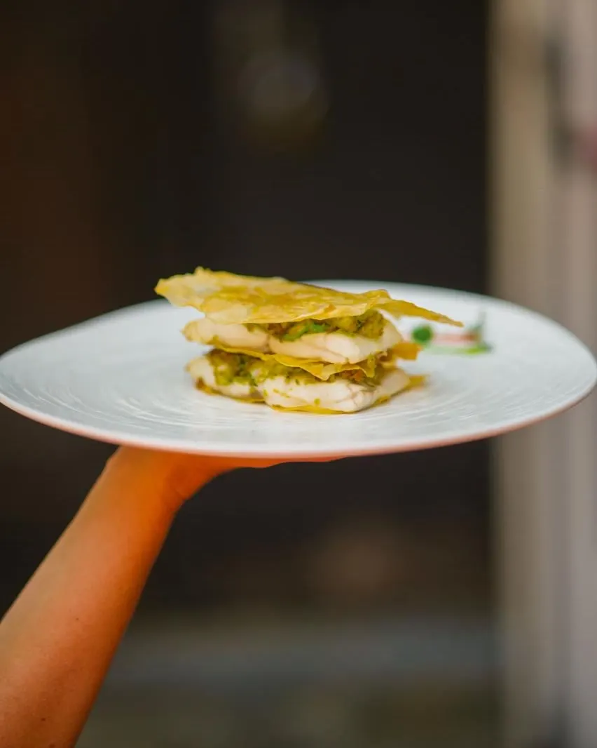 Filetti di pesce bianco a strati, alternati a sfoglie dorate croccanti e a un ripieno verde, su un piatto tenuto in mano
