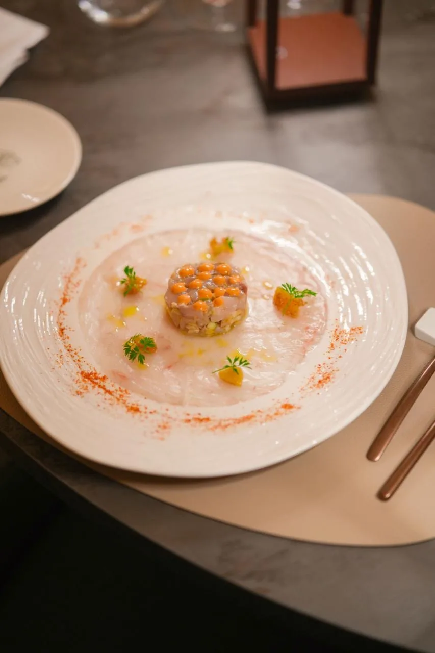 Tartare tonda su un carpaccio rosa, con perle arancioni sopra, ciuffetti verdi e polvere rossa sul bordo del piatto